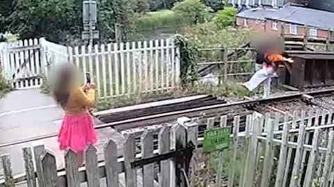 Network Rail CCTV footage of a railway line foot crossing. A person wearing a yellow top and pink skirt, with long brown hair is holding their phone up to take a photo of someone standing beside the railway line