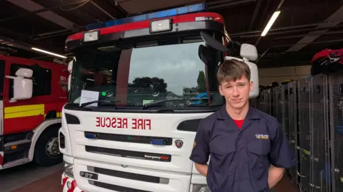 Lewis Owen has short brown hair. He is wearing a blue firefighter uniform and is standing in front of a white fire engine.