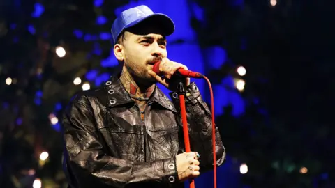 Zayn Malik wears a leather jacket and a blue hat. He is singing into a red microphone on a red microphone stand. 