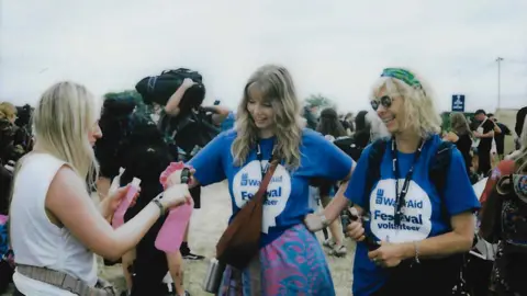 WaterAid A film photo showing two WaterAid volunteers wearing branded blue shirts, refilling a woman's pink water bottle while smiling.