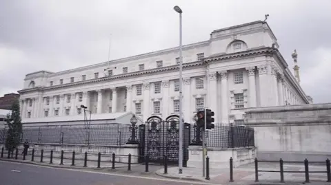 The High Court in Belfast. It is a white building with large back gates. It is situated on a main road beside a pedestrian crossing. 