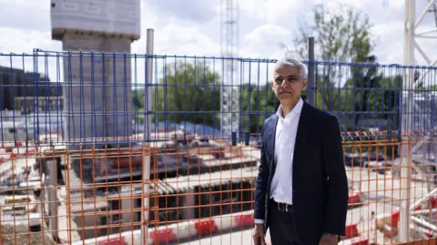 The mayor, wearing a dark suit and white shirt, stands in front of a building site. There are cranes and partially built houses and some trees in the background of the image. 