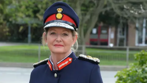 June Essex Susan Lousada, the Lord-Lieutenant of Bedfordshire, standing outside, with long blonde hair tied back, wearing a military cap, in red and blue colours, with a blue military jacket. She has pearl earrings in both ears. She is in front of a building and a bush is to the right. 