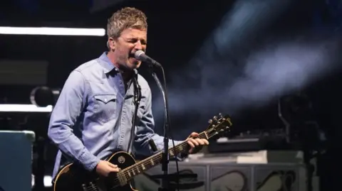 PA Media Noel Gallagher is singing into a microphone as Oasis takes to the stage at the Principality Stadium, Cardiff, for the band's long-awaited reunion tour kicks off in Wales.
