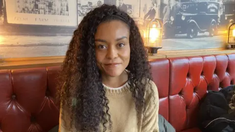 A woman with long curly brown hair and a pale brown top sitting on a red leather chair. Behind her are several archive photos of motor cars from bygone eras.