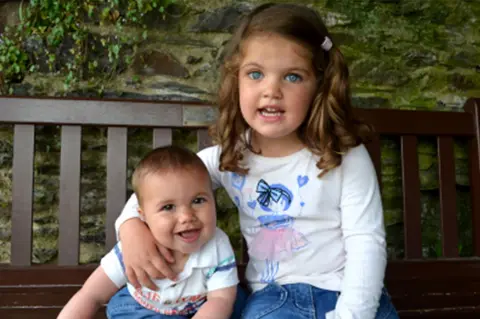 Family supplied James and Kate sit on a wooden bench outdoors. Kate has one arm around James and there is a stone wall and some greenery in the background.