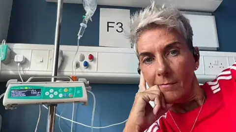 Carmel is looking at the camera and is leaning against her hand. She is in a hospital setting and wires and tubes can be seen beside her. She is wearing a red top with white stripes and has short grey hair. 