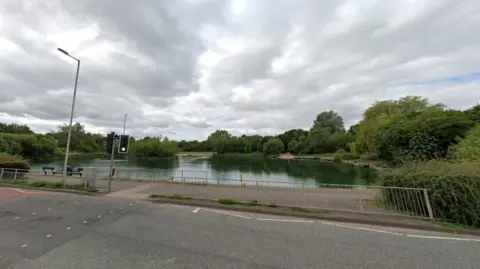 A body of water, behind a pedestrian crossing, is surrounded by trees. There is railing in the foreground and a streetlight.