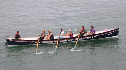 A long gig boat in a harbour with seven rowers on it. The boat is navy and has a white interior, as well as a white stripe and a red stripe towards the top of it. It has some words on the side which read 'StormFest 160 - Torbay RNLI - 22nd August 2026'. Six of the rowers are holding oars, which are light brown with teal coloured ends.