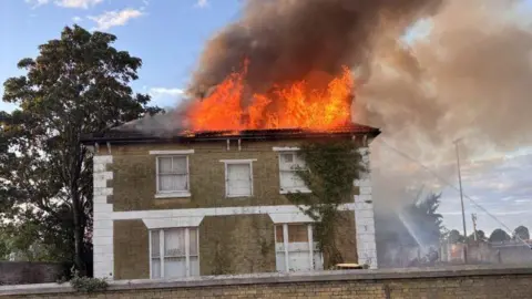 Large detached derelict building with flames coming from the roof. And water being sprayed onto the building from firefighters.