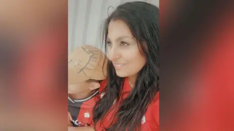 Nadiya Akhtar-Shahzad A woman with long black hair and her son wearing a brown cap watch a football match. Both are wearing red and black football shirts. 