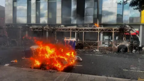 PA Media A car in the centre of the photo is fully engulfed in flames, while a shell of a bus - which had been burnt out - sits in the background.