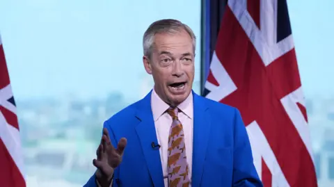 Gareth Fuller/PA Nigel Farage mid-way through giving a statement. He is wearing a light blue jacket, pink shirt and a tie with goldfish patterns on. A small microphone is pinned to his lapel. One hand is raised in a gesture to emphasise what he is saying. There are two large union flags behind him. 