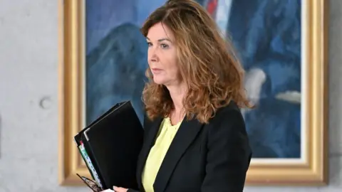 Getty Images Dorothy Bain, who has light brown curly hair, walks while holding a black folder. She is wearing a black suit jacket over a yellow top, and in front of a large painting with a gold frame hanging on a wall. 