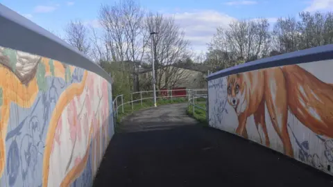 Kirsty Matheson A path over a bridge. The bridge has been painted on either side with a blue, yellow and white mural and there is a large picture of a fox on one side. 