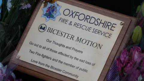 A gold plaque among the flower tributes. It includes the logos for Oxfordshire Fire and Rescue Service and Bicester Motion. It says: "Our thoughts and prayers go out to all of those affected by the sad loss of two fire-fighters and the member of the public/Love from the Bicester community"