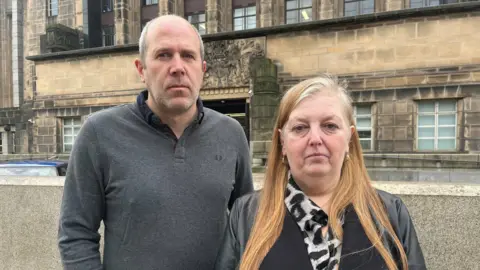 Alistair has short receding hair and stubble. He is wearing a grey jumper. Sharon has long red hair. She is wearing a black jacket.