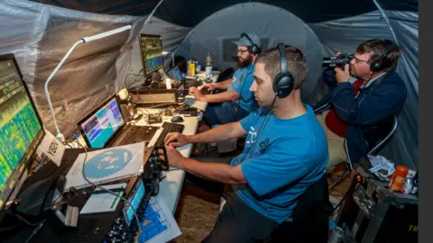 Radio Society of Great Britain Two men in blue tshirts sat at computer consoles with a range of electronic equipment in front of them and a man with a camera sat behind them