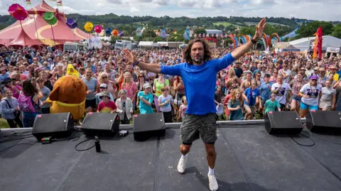 PA A man wearing a blue long t-shirt in front of a huge crowd of people while standing stage