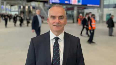 White short-haired man wearing a dark blue suit, white shirt and a navy tie with Translink patterned on it in turquoise standing infront of transport timetables at Grand Central Station