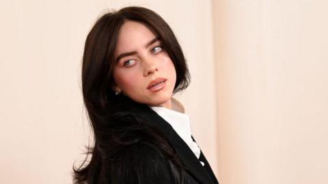 Billie Eilish at the Oscars in March 2024. Billie is a 22-year-old woman with long black hair and green eyes. She looks over her right shoulder, tilting her head back. She wears a black blazer over a white shirt. 