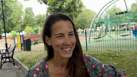 Emily Jennings stood in front of an outdoor play park. She is smiling and looking to the right, and is wearing a blue and pink patterned top. She has long brown hair.
