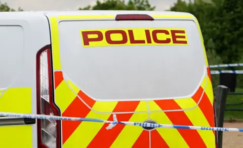 A generic picture of a white police van which has orange and yellow markings and a blue and white police tape in front of it. 
