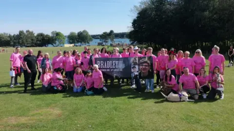 Group of charity walkers, dressed in pink, pose with a banner at Pitsford Reservoir on a sunny day