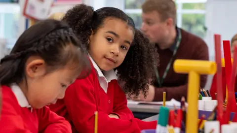 Getty Images A primary school aged girl in a red school uniform looks at the camera as she sits in class. Another student, also a girl in seen in the foreground looking down. A teacher talks to someone else in the background. 