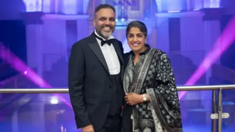 Dhinsa family A man and a woman stood on a balcony which has glass panels. They are wearing dark clothes and smiling at the camera. There are purple lights behind them. 