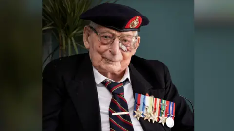 PA Jim Wren wearing a white collared shirt, black, red and gold striped tie, black blazer, a military cap and a row of colourful medals on his lapel. He is sitting in a chair, smiling at the camera. Behind him there is a potted plant and a dark teal wall.