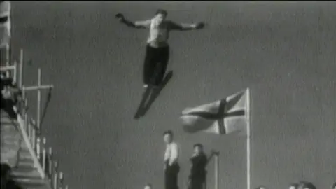 A black and white photo of a ski jumper taking off and flying through the air, with his arms outstretched. A Norwegian flag is fluttering on a flagpole behind him.