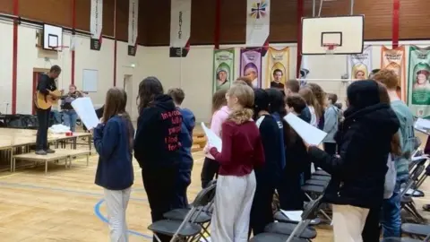 A photo of rows of children holding paper and singing. There are a mix of girls and boys and everyone is wearing comfortable jumpers and casual clothing. There is a woman standing on steps with a guitar.