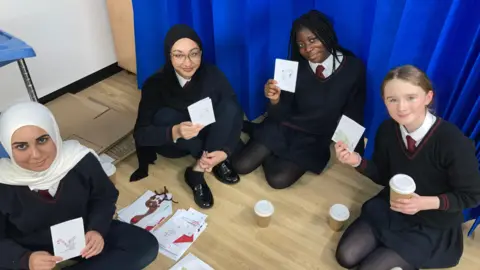 Four girls dressed in dark school uniforms are sitting on the laminate wooden floor of a hospital room with a blue curtain behind them. They are holding hand-made Christmas cards, with more on the floor between them. Two of the girls are wearing headscarves, one has black braided hair and one has light-brown hair tied back.