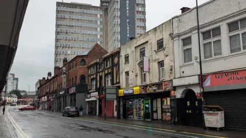 A general image of Bridge Street - an empty road with a row of shops and two high-rise buildings in the background.