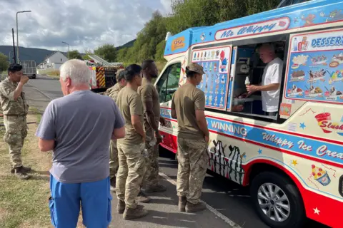Scott Emanuel A group of soldiers are stood in a queue towards an ice cream van. In the van is a man holding out an ice cream.