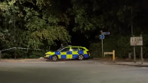 Police car and tape across a wooded area off a roundabout with a sign to Swaythling.