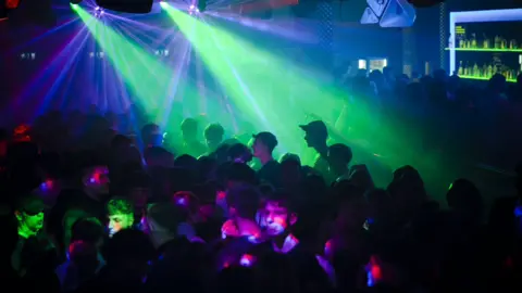 Revellers in a nightclub, silhouetted against brightly coloured lights in a dark venue. A bar is lit up in the background and speakers hang overhead.