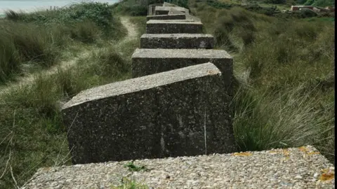 John Fairhall/BBC A row of concrete anti-tank defences. They are large rectangular grey blocks. They are sitting on a clifftop with grass behind them to the right and a sandy path and more grass in front of them to the left. On the horizon is a glimpse of the sea. 