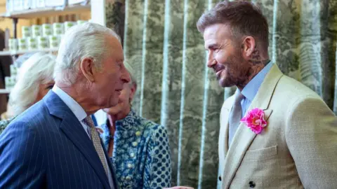 A man with grey hair and a blue suit is talking to a man with blight brown hair and a cream suit, with a bright pink flower in his lapel.