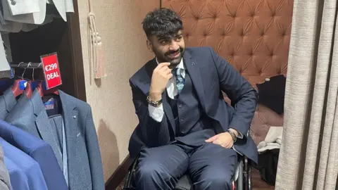 Yousaf Ghaffar A man sits in a wheelchair, smiling. The man has dark hair and facial hair and is wearing a navy suit with a blue tie and a white shirt. He is in a changing room booth and on his right there is a rack of suit jackets with a price tag above them. 