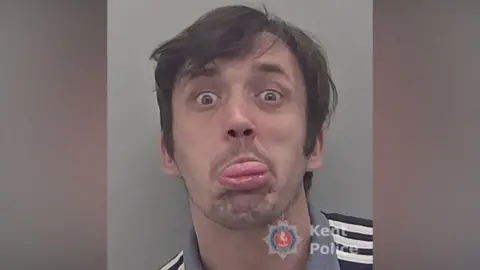 A mugshot of a man, with unkempt brown hair. He is sticking his tongue out.