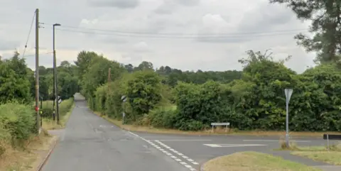 A road lined with trees and bushes. Another road, also lined with greenery, joins it at a junction.