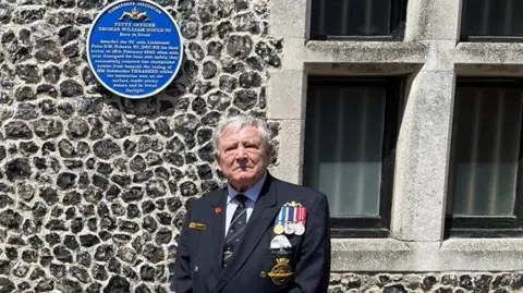 Veteran Sandy Powell is wearing medals and standing beside a blue commemorative plaque mounted on a stone wall outside a building.