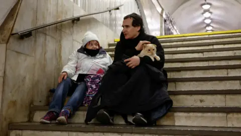 Reuters People take shelter inside a metro station during a Russian military attack. One man can be seen holding a cat while stroking its head while sitting next to a woman on the steps of a Metro station