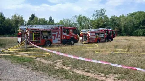 Fire engines are on a field that is cordoned off with red and white tape and long yellow hose reel jets are lying on the ground. There are trees around the edge of the field. The grass is yellow and dry.