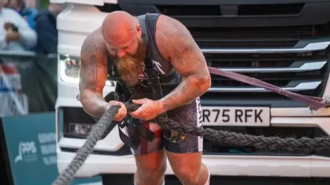 Ultimate Strongman A man in a dark leotard, with a bald head and long brown beard is pulling a lorry with a harness on his back. He is holding a thick black rope.