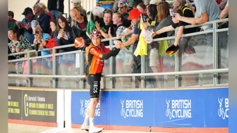 PA Media Thomas, wearing an orange top and black shorts, signs autographs for fans in the velodrome