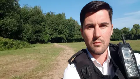 Handout photo issued by the Crown Prosecution Service of Ryan Connolly who is wearing a police uniform of white shirt and a black vest which has a communication device attached to it. He has straight dark hair, brushed into a side parting and has slight facial stubble. He looks directly into the camera, behind him a wide open space of grass is visible, with a path worn into it. Beyond that are trees in leaf and a bright blue sky