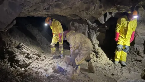 Three people in yellow cave suits can be seen in a muddy cave. One is digging and the other two are looking on.